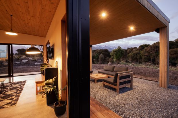 A warm, modern indoor-outdoor living space viewed through a large dark-framed opening. The interior has a timber-lined ceiling, polished concrete floor, pendant lighting, potted plants, and a patterned rug, while the covered outdoor area features a timber ceiling and posts, timber-framed lounge seating, and recessed lighting overlooking a natural landscape.