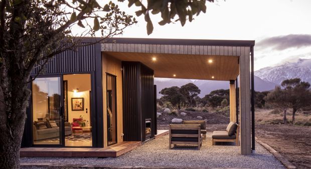 A compact modern cabin or pavilion set in a rugged alpine landscape. The dark vertically ribbed metal-clad structure has a simple flat roof and a large covered outdoor living area framed by timber-lined soffits and exposed steel supports. Floor-to-ceiling glazing reveals a warm living room inside, while outdoor seating occupies the gravel terrace. Mountain terrain and native vegetation are visible beyond.