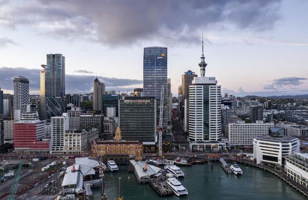 A wide aerial view of a dense waterfront city centre, showing a harbour with ferry terminals and boats in the foreground, surrounded by modern high-rise commercial buildings and Auckland's Sky Tower in the background under a cloudy sky.