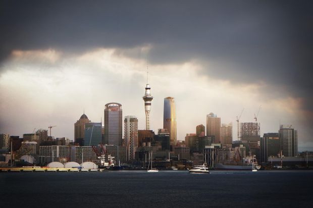A wide waterfront view of a dense modern city skyline under dramatic storm clouds, with high-rise office and residential buildings, a prominent communications tower, construction cranes, and boats on the water in the foreground.