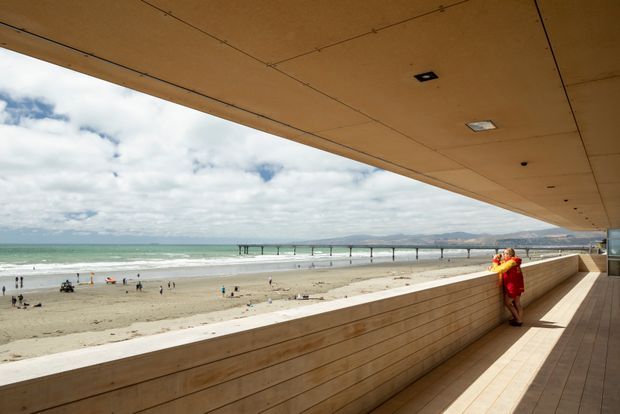 A wide covered beachfront terrace or balcony overlooking a sandy beach, ocean, and a long pier. The space features a broad timber-clad overhanging ceiling, horizontal timber wall cladding and a timber deck, with a low viewing wall and recessed ceiling lights. People stand at the edge looking toward the beach and distant coastline.