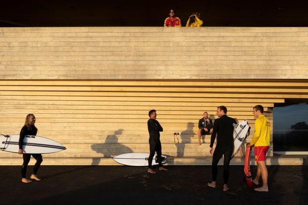 Exterior architectural photograph of a modern timber-clad surf club or beach facility. A broad horizontal timber facade forms the main subject, with a deep dark overhang and an upper-level balcony or opening. Surfers carrying boards gather on the dark paved ground in front of the building, and a large opening on the right reveals a darker interior or covered area.