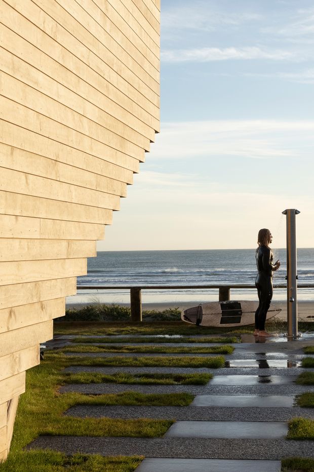 A contemporary coastal building exterior beside a beach, featuring a prominent projecting facade clad in horizontal light timber boards. A wet paved walkway crosses a landscaped lawn toward an outdoor rinse shower and railing, with the ocean visible beyond. A surfer in a wetsuit stands near the shower holding a surfboard.