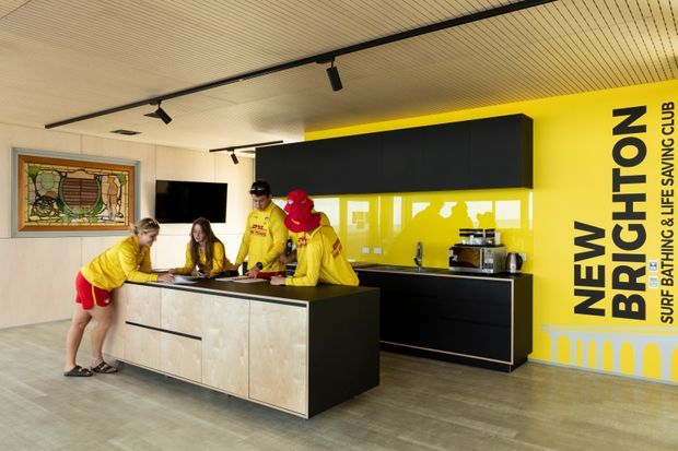 A brightly branded commercial kitchen or hospitality space with a large central island, black and pale timber cabinetry, a vivid yellow glass splashback and feature wall, open shelving, sink, and coffee equipment. Track lighting runs across a light linear-panel ceiling, while several people in yellow uniforms gather around the island. A framed stained-glass artwork and wall-mounted screen are visible on the opposite wall.