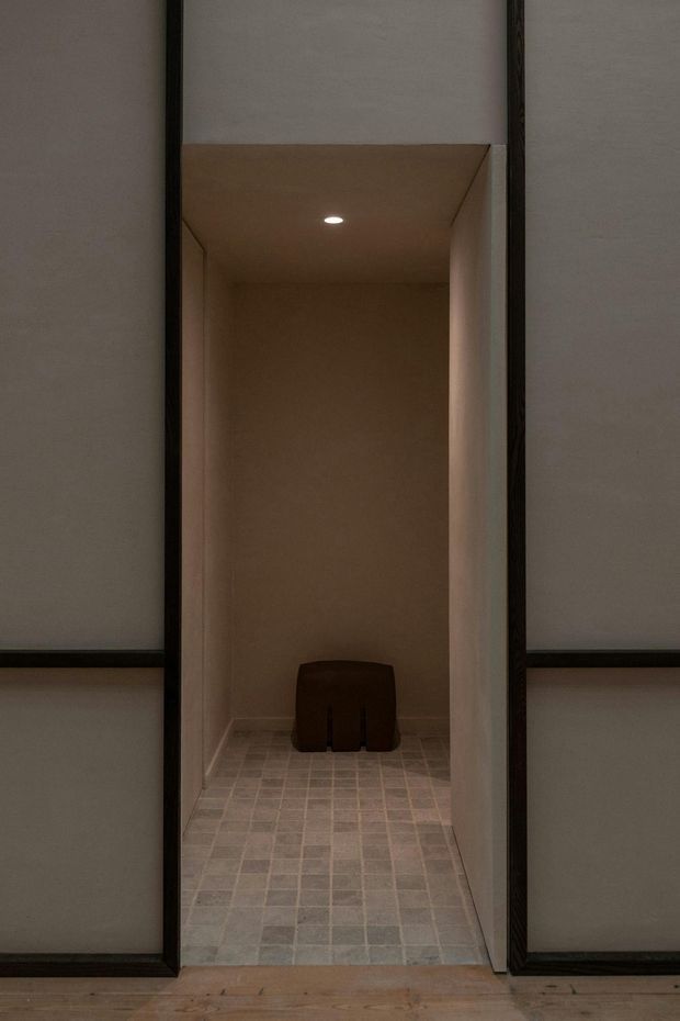 A narrow, minimalist tiled alcove or small bathroom space viewed through a dark-framed opening. The room has warm beige plastered walls, a small recessed ceiling light, pale square tiled flooring, and a dark low stool or compact fixture at the rear. Timber flooring and large dark-framed wall panels surround the opening.