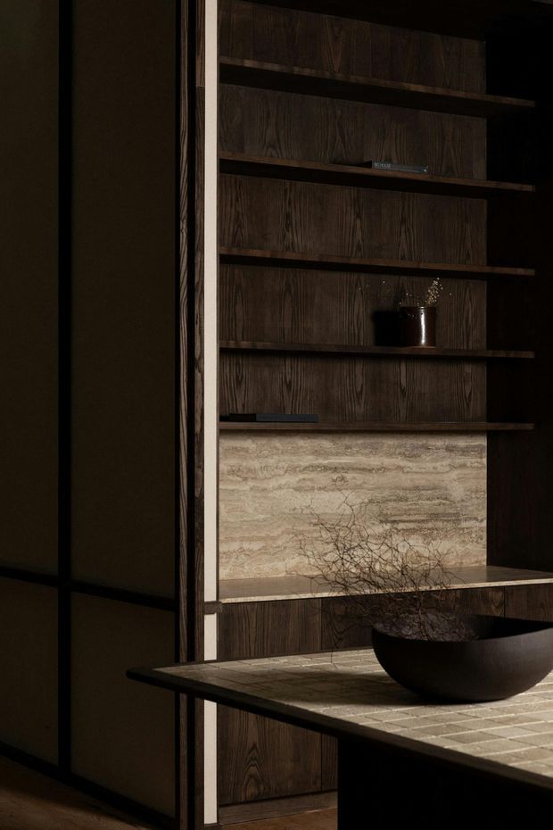 A moody, refined interior vignette centred on a dark timber built-in shelving wall with multiple open shelves, a pale stone feature panel, and lower cabinetry. In the foreground is a substantial island or console with a textured light stone or tile surface, holding a large dark bowl with branches. The space is dimly lit with strong contrast, rich wood tones, and minimalist decorative styling.