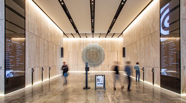 A spacious contemporary commercial lobby or reception area with tall light timber wall panels, polished stone flooring, illuminated edges, integrated linear ceiling lighting, large digital information displays, and a prominent circular sculptural feature wall. Several visitors and a freestanding information sign are visible.
