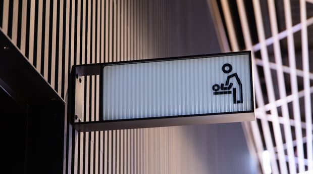 Close-up of a contemporary architectural wayfinding sign projecting from a wall, with a black metal frame, translucent ribbed panel, and a black accessibility symbol. Vertical slatted wall detailing and geometric glazing or screening are visible in the background.