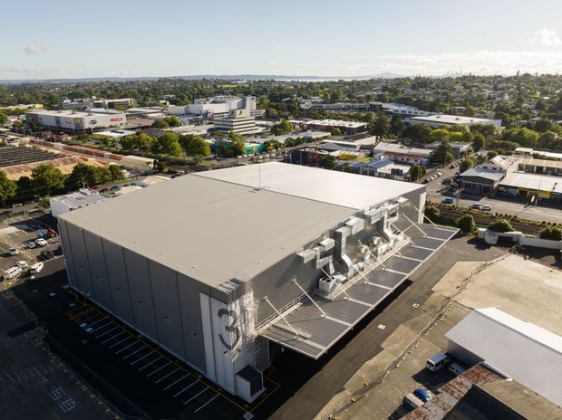 Aerial view of a large modern industrial or commercial warehouse building with a broad low-pitched roof, tall grey metal-clad walls, rooftop mechanical equipment, loading or service areas, and marked parking around it. The building sits within a dense suburban commercial and light-industrial district, with low-rise buildings and trees extending toward the horizon.