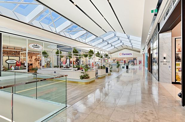 Bright modern indoor shopping mall corridor with polished stone flooring, glass balustrades, retail storefronts, planted display areas, and extensive skylights bringing in natural light. Visible shops include Farmers and fashion retailers, with a broad open circulation space and contemporary linear ceiling details.