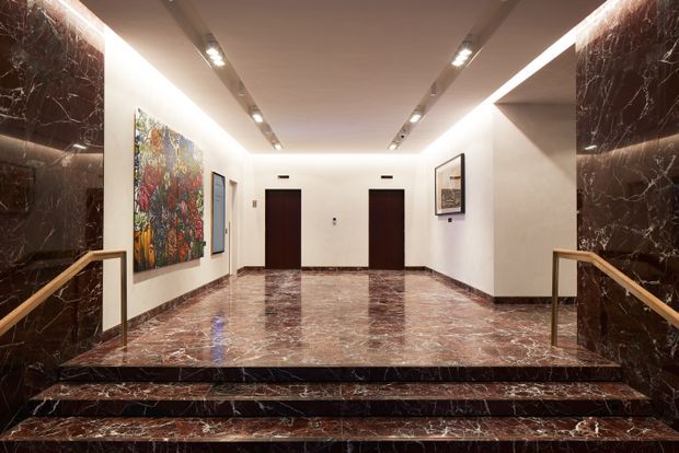 A polished interior corridor or lift lobby with two dark timber doors, a short flight of marble-clad steps, marble wall bases and columns, brass-toned handrails, recessed linear lighting, and framed artwork. The space has a formal, refined hotel or commercial character with strong symmetry and glossy stone surfaces.