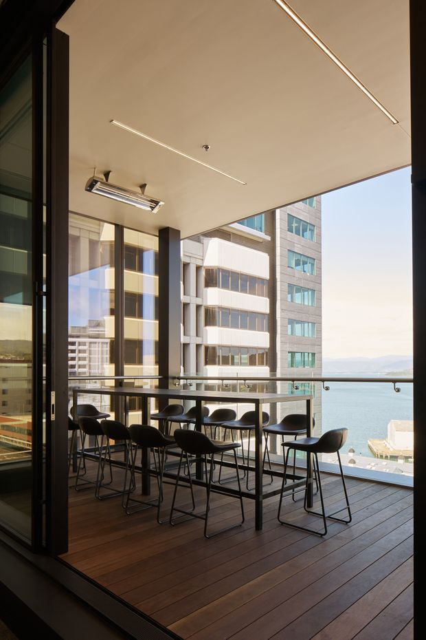 A covered high-rise balcony or terrace with expansive glazing and a glass balustrade, furnished with a long narrow bar-height table and black stools. Dark timber decking, slim linear ceiling lights, and dark metal framing create a contemporary hospitality-style outdoor seating area. Modern commercial buildings and a waterfront view are visible beyond.