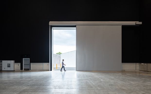 A large, dark industrial-style interior with a broad polished concrete floor and a tall open loading-bay doorway. Minimal built-in utility elements line the walls, while a person provides scale against the expansive space. Daylight enters through the opening, contrasting with the black walls and ceiling.