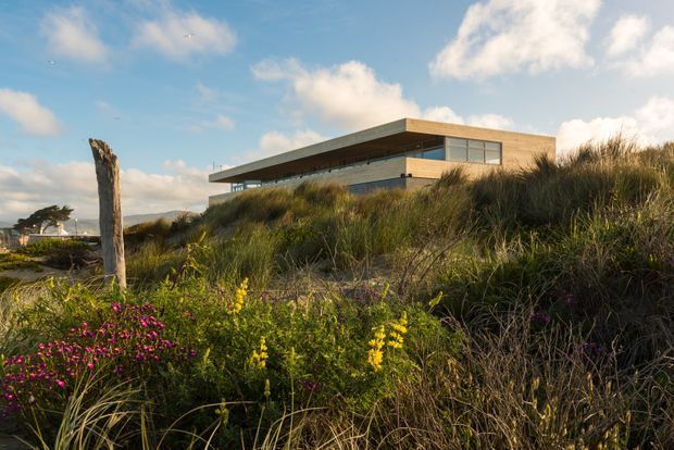 A contemporary, low-profile flat-roofed house set among coastal sand dunes and native grasses. The building features extensive horizontal glazing, warm timber-clad upper walls, projecting roof planes and a long cantilevered form. Wild coastal planting dominates the foreground, with soft late-day light and blue sky creating a natural seaside atmosphere.