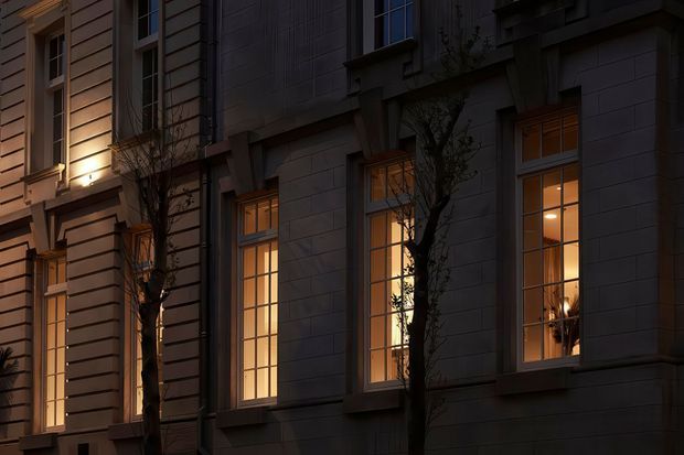 A close-up dusk view of a refined multi-storey building facade with tall, warm-lit rectangular windows, dark painted masonry or rendered wall panels, projecting window surrounds, and classical vertical detailing. Narrow street trees partially obscure the facade, while the illuminated interiors create a warm contrast against the dark exterior.