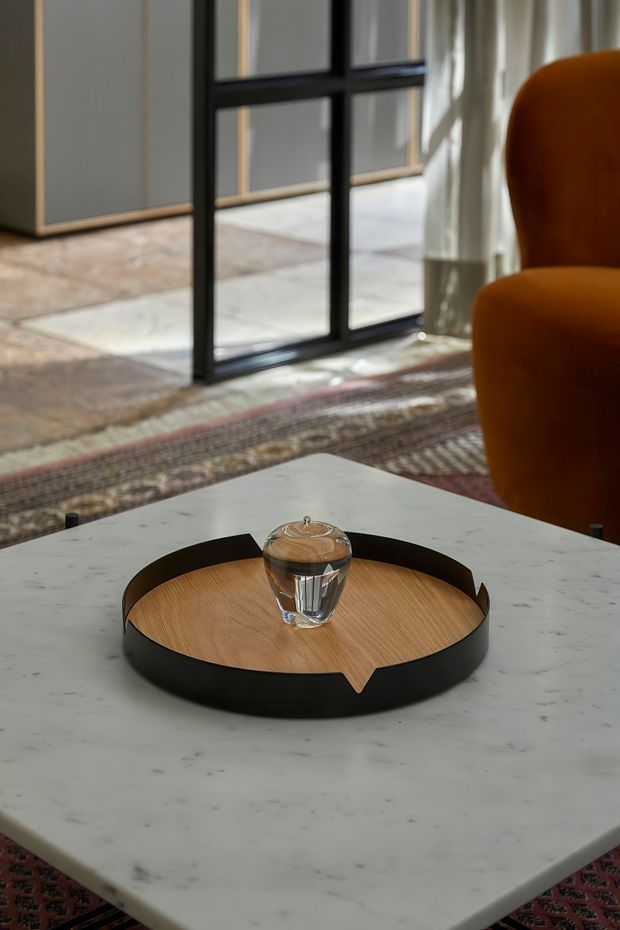 A close-up interior vignette showing a round white marble coffee table with a black metal and wood serving tray holding a glass. An orange upholstered armchair, patterned rug, sheer curtains, and a black-framed glazed partition or doorway are visible in the softly lit background.