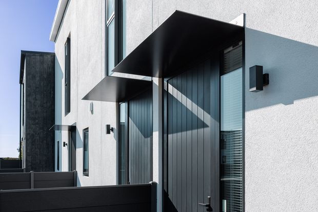 Exterior view of a contemporary multi-level residential facade with white textured rendered walls, tall narrow glazing, dark vertical cladding panels, and projecting black metal awnings over entry openings. A dark privacy screen or fence occupies the foreground, emphasizing the building's crisp geometric composition.