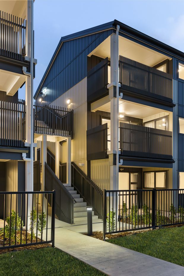 A contemporary multi-storey residential building photographed at dusk, featuring dark vertical metal cladding, repeated projecting balconies with black vertical balustrades, white structural posts and downpipes, and externally accessed stairs. A concrete pedestrian path leads through landscaped lawn and low planting to the entry areas, with warm interior and exterior lighting creating a refined evening presentation.