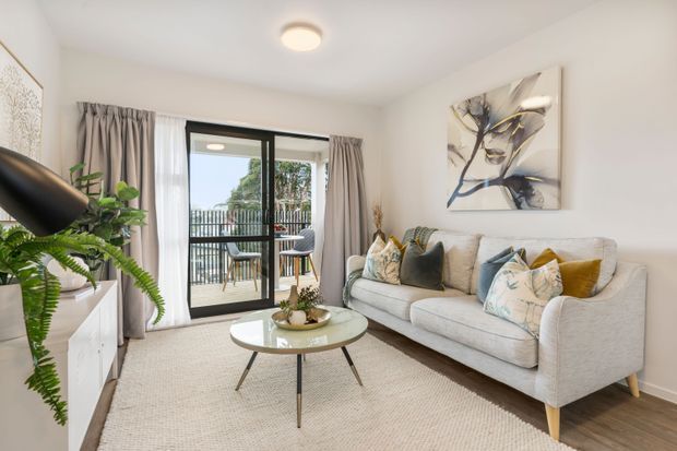 Bright, inviting contemporary living room with a light upholstered sofa, patterned and mustard accent cushions, round glass coffee table, pale textured rug, white walls, timber flooring, and soft grey curtains. A black-framed glazed door opens to a balcony, while indoor plants and abstract artwork add natural and decorative touches.