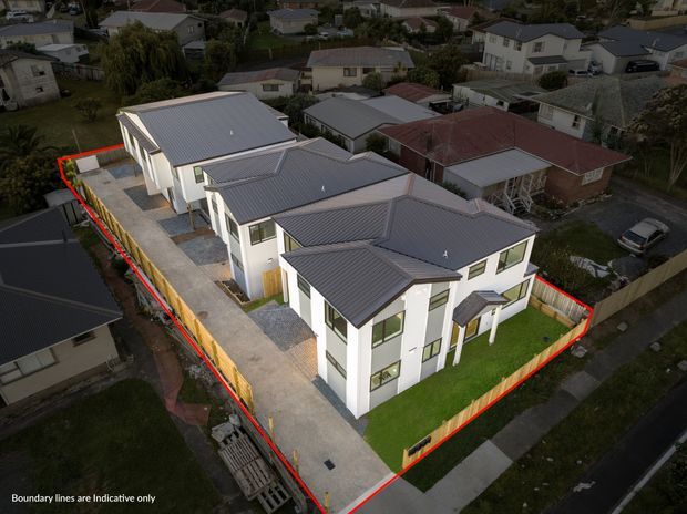 Aerial oblique view of a newly developed multi-unit residential property on a compact suburban site. Three attached, modern two-storey dwellings feature crisp white façades, dark grey metal gable roofs, large windows, private lawns, concrete driveways and timber boundary fencing. Red boundary lines outline the site, with surrounding suburban homes visible beyond.