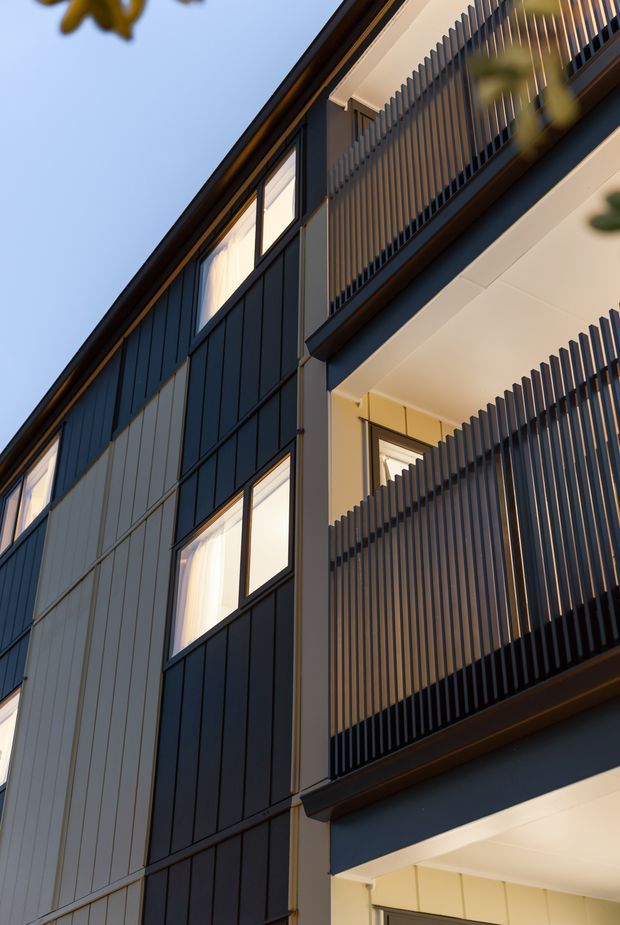 Upward-looking architectural photograph of a contemporary multi-storey building facade. The facade combines dark vertical cladding, light panel sections, large glazed windows, and projecting balconies screened by closely spaced vertical slats. Warm interior lighting contrasts with the cool blue-grey exterior, while foliage is softly visible at the edges.