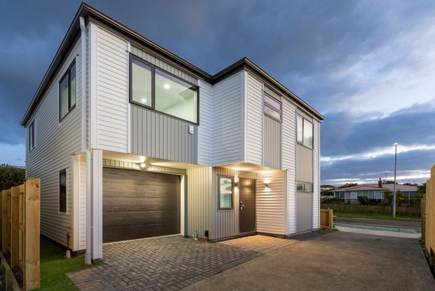 Exterior view of a contemporary multi-storey residence at dusk, featuring light horizontal weatherboard cladding, darker vertical cladding accents, projecting box-like upper volumes, large windows, an integrated single garage, and a paved driveway leading to the entry.