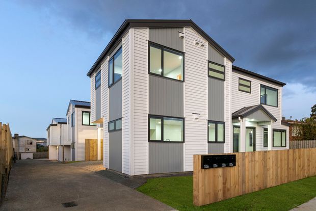 Exterior view of a contemporary multi-storey townhouse development with white horizontal cladding, dark grey vertical feature panels, dark-framed windows, pitched roof forms, and timber privacy fencing. A paved driveway runs alongside the homes, with small landscaped lawns at the front.