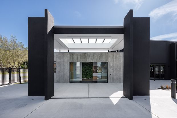 Front-facing exterior view of a contemporary commercial or institutional entrance framed by tall dark vertical architectural fins. The entry features a raw grey concrete wall, broad glazed sliding doors, a projecting canopy with a large rectangular skylight, and a clean concrete forecourt. The composition is geometric and minimalist, with adjacent dark-clad building volumes visible to the right.