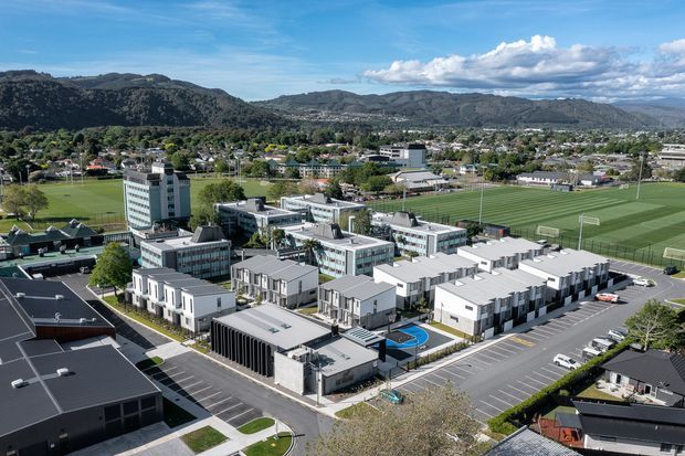 Aerial view of a contemporary multi-building institutional or commercial complex set within an urban suburban neighbourhood. The development features flat-roofed white and grey buildings, extensive parking, landscaped paths, a small blue sports court, and large adjacent playing fields, with wooded hills in the background.