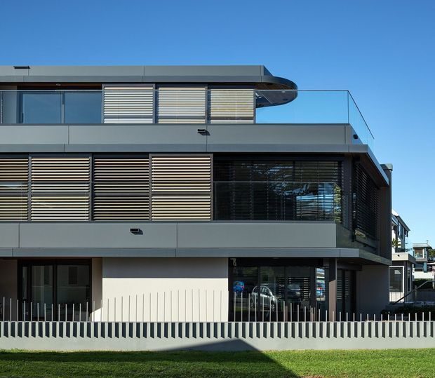 Contemporary multi-storey residential building viewed from the street, with a dark grey and white facade, extensive glazing, horizontal external timber-look louvres, projecting balconies, and a distinctive rounded roof edge. A low vertical-slat boundary fence and lawn occupy the foreground under a clear blue sky.