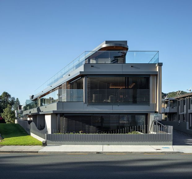 Street-facing exterior view of a contemporary multi-level residential building with a strongly horizontal composition, dark grey rendered or metal-clad walls, extensive glazing screened by horizontal louvres, glass balustrades, and a rooftop terrace or pavilion. The building sits on a gently sloping suburban site with a small lawn, driveway access, and neighbouring homes visible to the side.