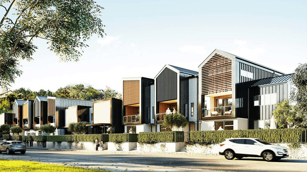 Street-facing view of a contemporary multi-unit residential development composed of several narrow, vertically articulated dwellings. The buildings feature steep gabled roof forms, dark metal cladding, pale rendered walls, timber-toned vertical screens and horizontal timber battens, with balconies, large windows and landscaped front gardens along the road.