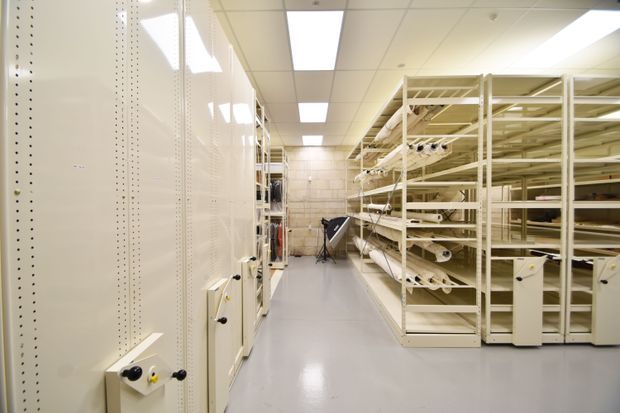 A bright, utilitarian storage room with floor-to-ceiling cream metal shelving and compact storage cabinets arranged along both sides of a central aisle. Rolled materials and equipment are stored on the open shelving. The space has smooth grey flooring, pale walls, and a suspended acoustic ceiling with rectangular fluorescent light panels.