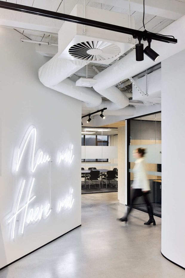 A bright contemporary commercial office interior with a polished concrete floor, white curved partition walls, exposed white-painted services and large ducts across the ceiling, black track lighting, and glass meeting-room partitions. A glowing script neon sign forms a feature on the left wall, while a blurred person walks through the corridor. A meeting room with a table and chairs is visible beyond the entry passage.
