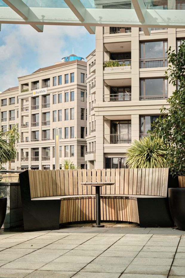 A sunny urban apartment or commercial courtyard terrace viewed from a paved outdoor seating area. The foreground contains curved timber-slat bench seating, a small round pedestal table, large black planters, and a translucent glass-and-timber overhead canopy. Mid-rise contemporary apartment buildings with balconies and light beige rendered facades surround the space, with landscaped planting adding greenery.