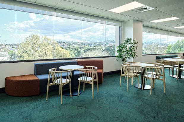 A bright commercial lounge and breakout area with floor-to-ceiling windows overlooking a leafy urban landscape. The space features teal carpet, low modular upholstered seating in navy, rust and brown, and several small round tables with light timber or painted wood chairs. A potted plant, suspended ceiling, and recessed fluorescent lighting complete the contemporary office fit-out.