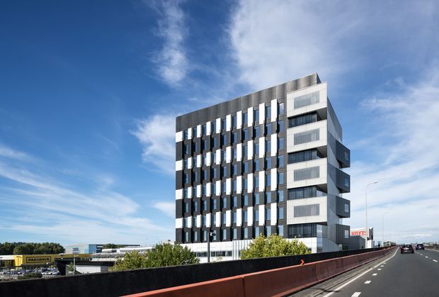 Exterior architectural photograph of a contemporary multi-storey commercial building beside a motorway. The facade combines dark vertical panels, regularly spaced projecting fins, extensive glazing, and protruding white-framed window bays. The building sits in an urban commercial setting, with road infrastructure and low-rise development around it under a bright blue sky.