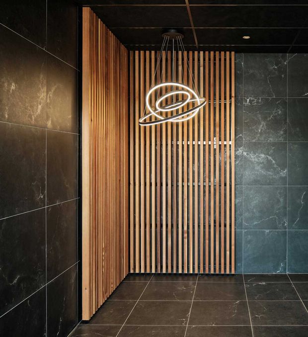 A dark, refined interior featuring large-format charcoal stone-look wall and floor tiles, warm vertical timber battens forming a screened partition, and a sculptural illuminated ring pendant light. The space appears to be a contemporary bathroom or wet-area entry, though sanitary fixtures are not visible.