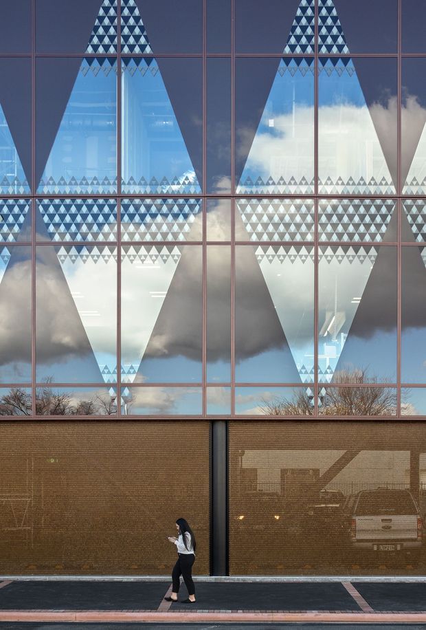 Street-level view of a contemporary commercial building facade dominated by a large gridded glazed curtain wall. The glazing reflects sky, clouds, trees, and surrounding architecture, while repeated triangular geometric patterns create a bold graphic effect across the upper windows. The lower facade is clad in warm brown textured brick or fine masonry, with a pedestrian walking along the pavement in front.