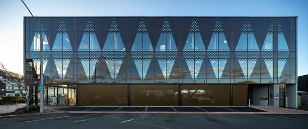 Street-facing exterior view of a contemporary multi-storey commercial building. The upper facade is enclosed in dark-framed glazing with a striking repeating geometric triangular pattern, while the ground level has recessed glazed entrances and dark solid panels. The building sits directly along a paved urban frontage and is photographed in daylight.