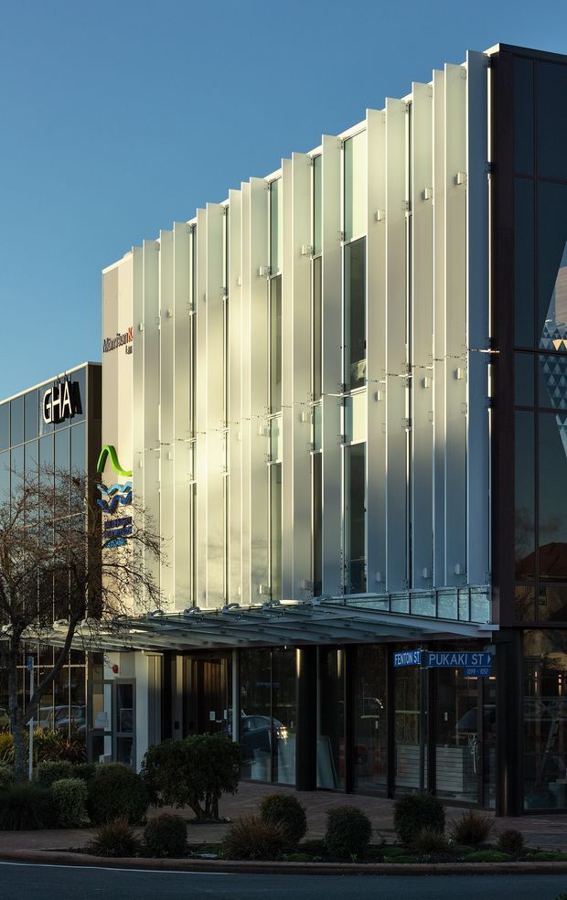 Exterior street-level view of a contemporary multi-storey commercial building with a strongly articulated pale metal facade made from repeated vertical fins, glazed entrances, and a dark glazed side volume. The building fronts a landscaped urban streetscape with signage and street-name signs visible near the entrance.