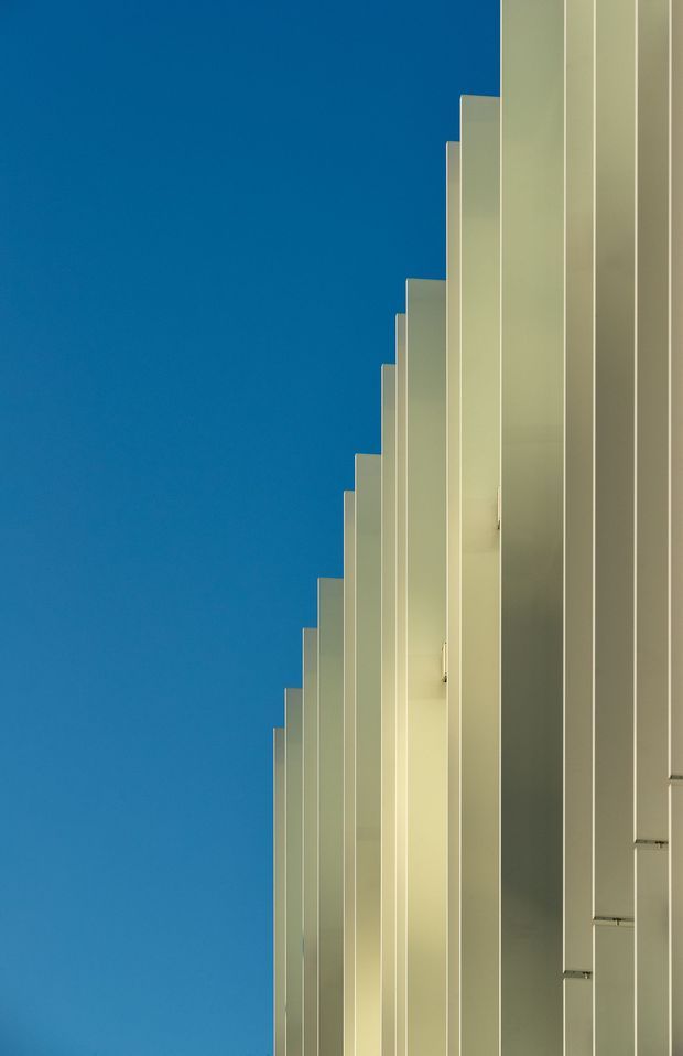 Minimalist architectural exterior detail showing a rhythmic series of tall, pale vertical facade fins or panels receding diagonally upward against a clear deep-blue sky. The composition emphasizes repetition, clean geometry, strong vertical lines, and contrasting light and shadow, with no visible interior or landscaping.