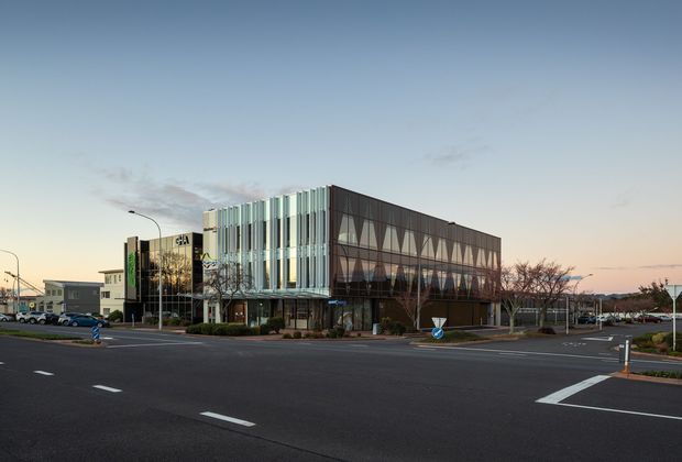 A contemporary multi-storey commercial building viewed from a broad urban road intersection at dusk. The facade combines vertical pale fins and glazing on one side with a dark geometric patterned facade on the other, creating a strong angular corner expression. Low landscaping and adjacent commercial buildings surround the site; the image is dominated by the building exterior and streetscape.