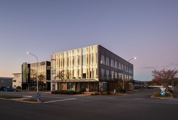 A contemporary multi-storey commercial building photographed at dusk from a street intersection. The prominent facade combines extensive glazed curtain walling with vertical illuminated fins and darker geometric side panels. Landscaped pedestrian edges, street lighting, road markings, and neighbouring low-rise buildings surround the property.