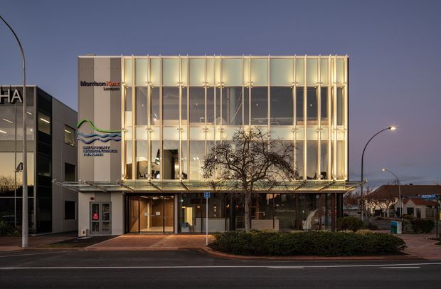 Twilight street-level photograph of a contemporary multi-storey commercial building. The facade combines extensive curtain-wall glazing with a prominent grid of slim vertical fins and warm linear illumination. A glazed canopy shelters the recessed ground-floor entry, with commercial signage and neighbouring retail buildings visible around the site.