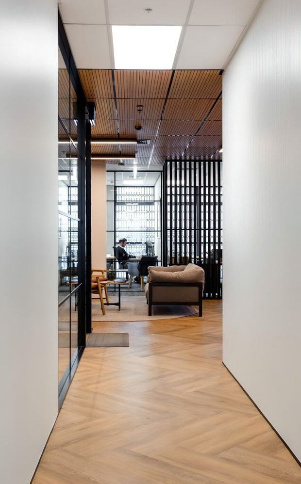 A contemporary office interior viewed from a narrow corridor, featuring warm herringbone timber flooring, white walls, black-framed glazing, a timber slatted ceiling, and a black vertical screen partition. A lounge chair and small table sit near a workstation in the background.
