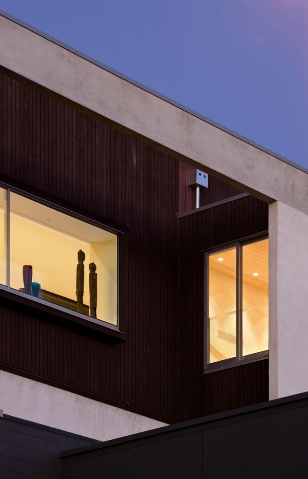 Upward-looking exterior architectural photograph of a contemporary multi-level building at dusk. The facade combines dark vertically ribbed timber cladding with pale rendered or concrete bands, crisp flat rooflines, and large illuminated windows. A recessed courtyard-like opening and projecting geometric volumes create a strong modern composition.