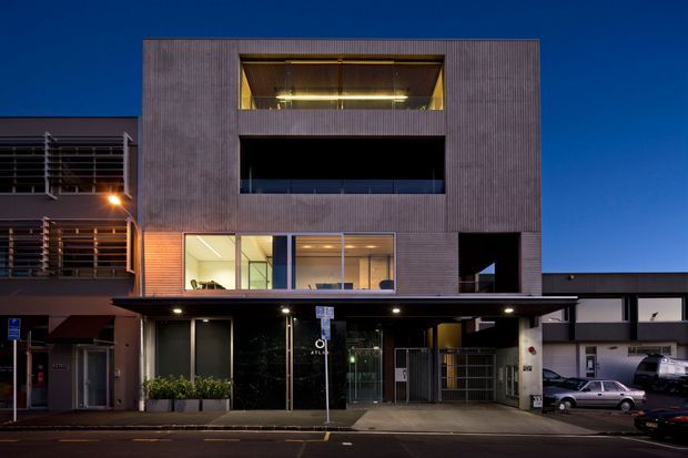 A dusk exterior photograph of a contemporary multi-storey urban building with a rectilinear, box-like form. The dark textured facade includes recessed balconies and large illuminated windows, with a glazed ground-floor entry or commercial frontage, a projecting canopy, and an integrated vehicle access area. Adjacent buildings and parked cars are visible in the streetscape.