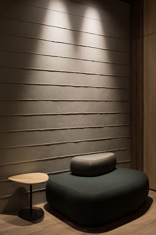 A moody, intimate interior vignette featuring a low, rounded dark upholstered lounge seat or ottoman with a matching cushion, positioned against a horizontally textured plaster wall. A small light timber side table with a black pedestal base sits alongside it, while vertical timber panelling frames the right side. Warm directional lighting creates strong shadows and highlights the tactile wall finish.