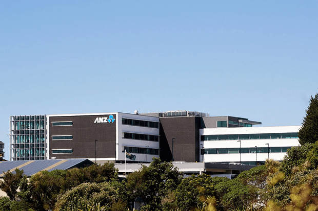 Exterior photograph of a multi-storey contemporary commercial office building set behind landscaped vegetation. The complex has dark masonry-clad volumes, extensive horizontal strip windows, glazed curtain-wall sections, projecting flat roof forms, and a visible ANZ logo.